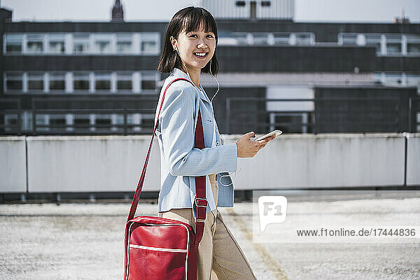 Smiling teenage girl wearing shoulder bag holding smart phone while walking on rooftop