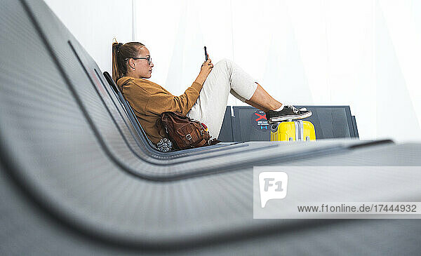 Frau benutzt Mobiltelefon  während sie auf Bank im Flughafen sitzt