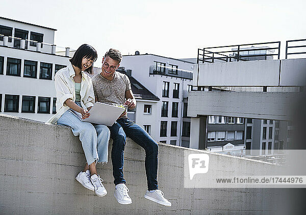 Smiling male and female friends using laptop while sitting on retaining wall