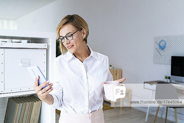Smiling businesswoman holding coffee mug while text messaging through mobile phone in office