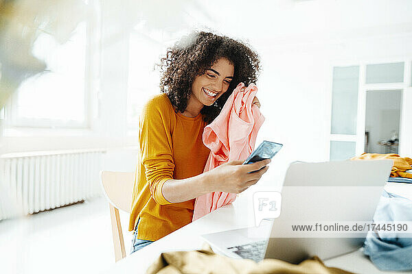 Happy woman with clothes using smart phone at home