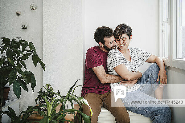Mid adult man embracing woman while sitting near window at home