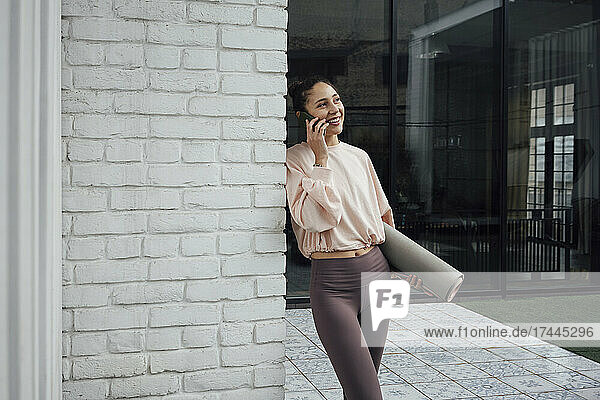 Smiling woman with exercise mat talking on mobile phone at terrace