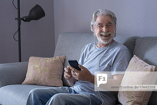 Happy man with mobile phone and in-ear headphones sitting at home