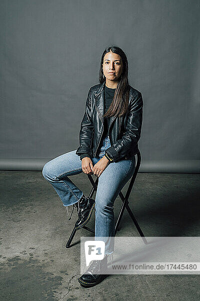 Young confident woman in leather jacket sitting on chair in studio