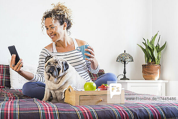 Woman using mobile phone while sitting with pug on bed