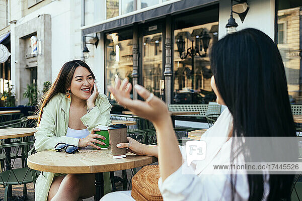 Female friends with coffee cup talking while sitting at sidewalk cafe
