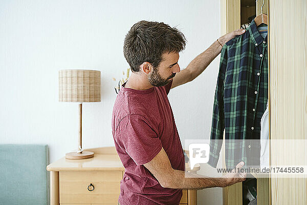 Mid adult man holding shirt while standing near closet at home