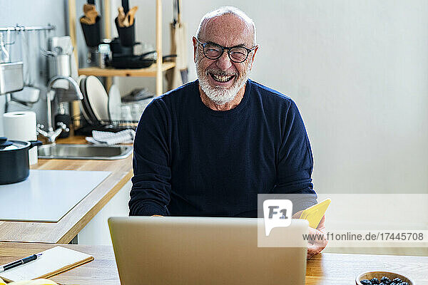 Cheerful senior man with mobile phone and laptop sitting in kitchen