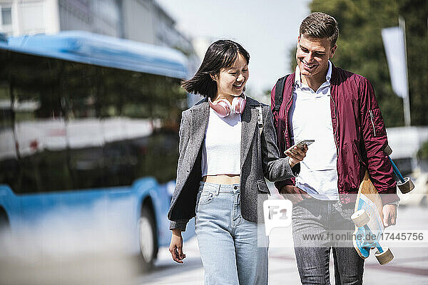 Smiling male and female friends sharing mobile phone while walking during sunny day