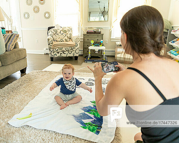 Baby boy sitting on blanket smiling as mother takes photograph.