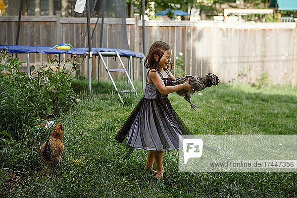 A little laughing girl holds a chicken and twirls in backyard garden
