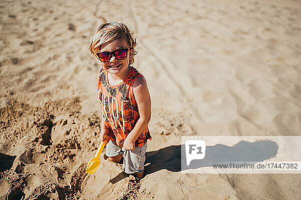 Happy young boy wearing sunglasses at beach playing with shovel