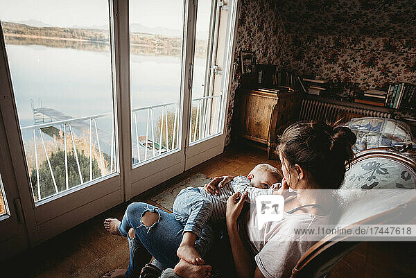 Mother breastfeeding baby on chair in vintage room looking out on lake