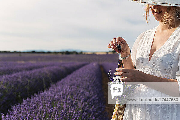 Unrecognizable woman in white dress holding a bottle of lavender oil.