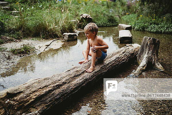 Young boy sitting on log in water in summer looking thoughtful