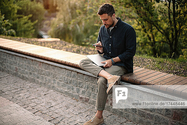 Man using smart phone and holding laptop while sitting on bench