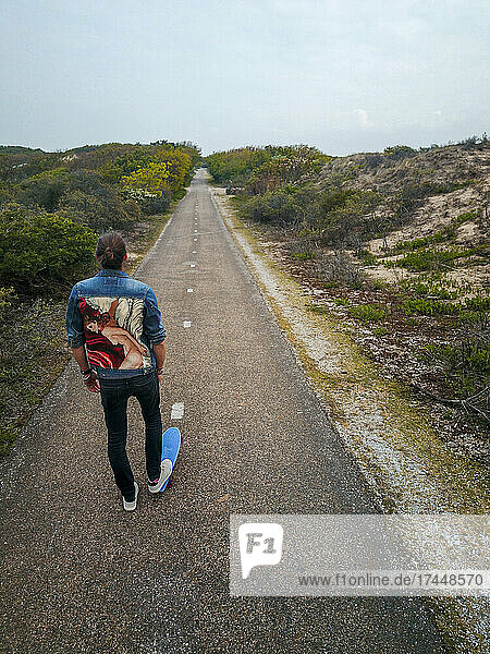A man and his skateboard facing a long deserted straight road