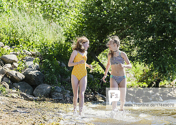Two happy tween teen girls in swimsuits running on lake shore.