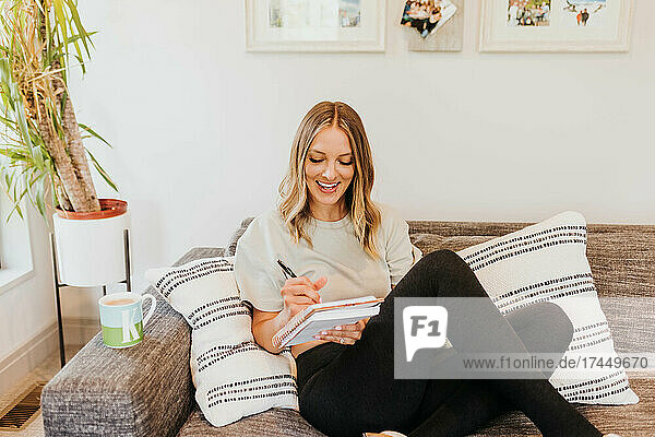 Wide shot of smiling woman sitting on couch writing in notebook