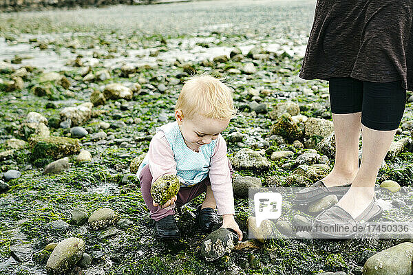A young girl crouched on a rocky beach next to her mom