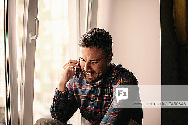Happy man talking on smart phone sitting by window at home