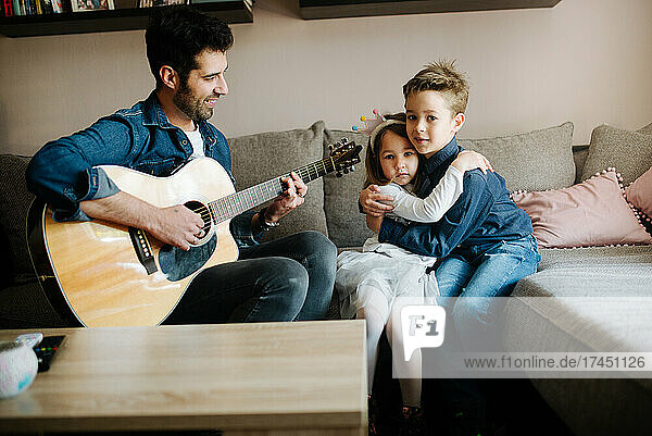 The father plays an acoustic guitar for the children at home.