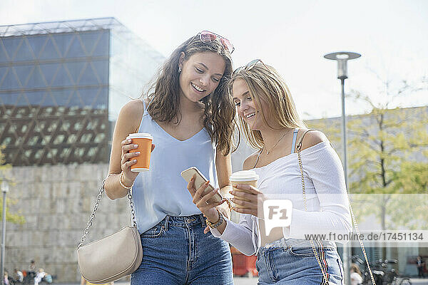 Two young women with coffee cups watching on mobilphone in City
