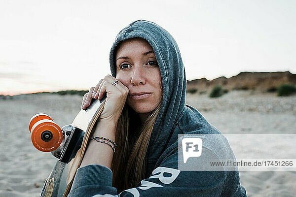 woman sat on the beach with her hood up with her skateboard