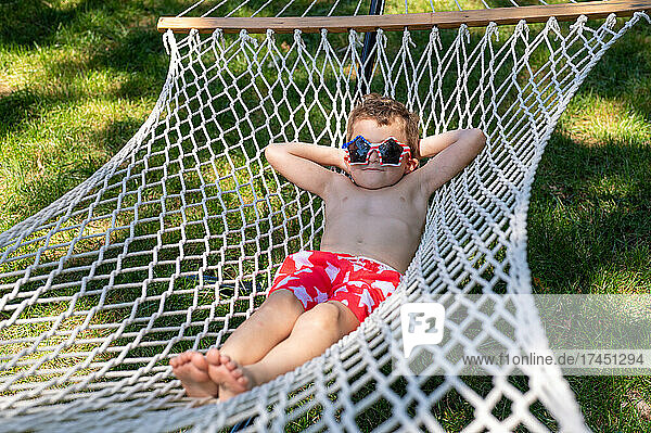 Silly boy relaxing on hammock in swimsuit and sunglasses