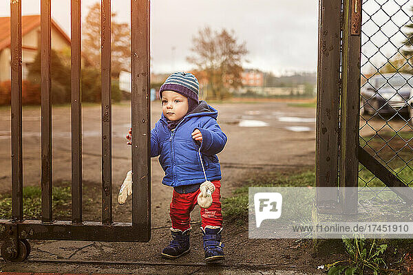 Small boy walking through gate wearing blue jacket and red trous