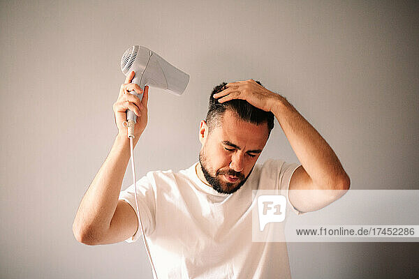 Man blow drying hair against gray wall at home