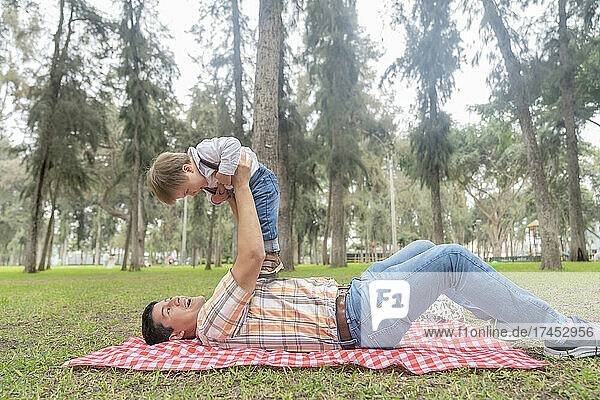 Man lying on a park as he raise his son with his arms