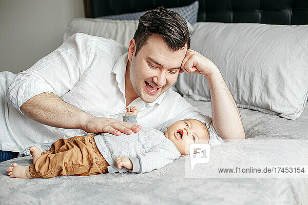 Happy proud dad lying on bed playing with newborn baby boy