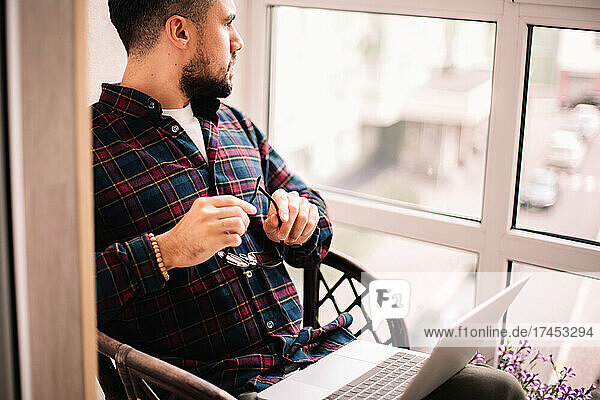 Thoughtful man looking through window working with laptop at home