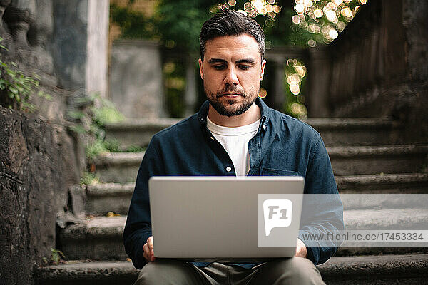 Serious man using laptop computer sitting on steps in city