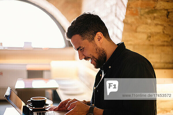 Happy man laughing while using laptop computer at cafe