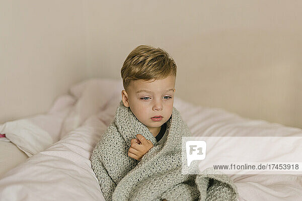 Ill and tired boy wrapped in grey blanket sitting on bed