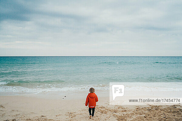 Back view of child wearing jacket walking to sea on cloudy day