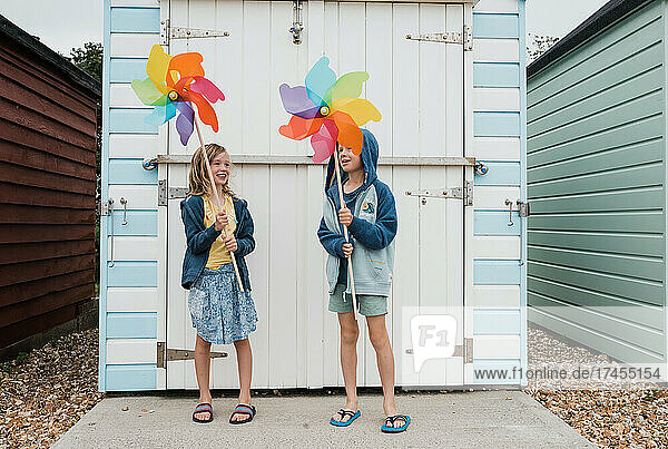siblings laughing and playing with windmills by beach huts