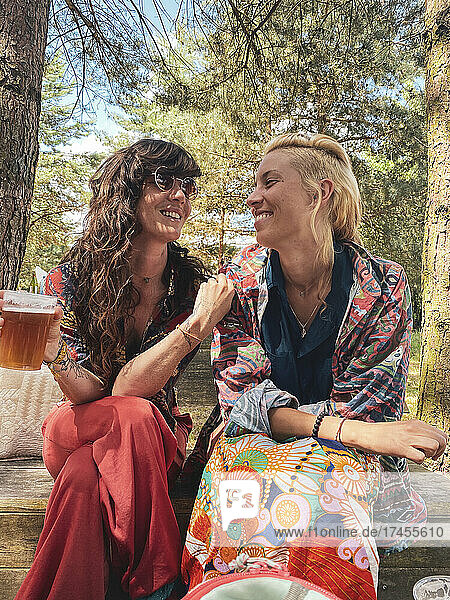 Happy queer women couple smile affectionately over beer in forrest