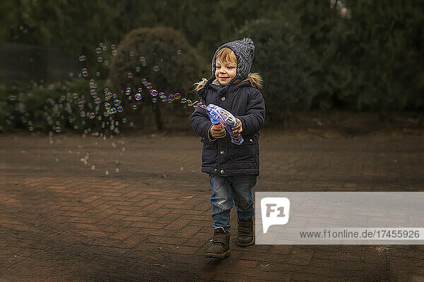 Toddler in warm clothes playing with bubble gun outside