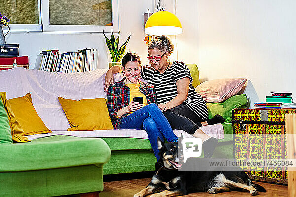 Queer couple cuddling on yellow couch at home with flowers and books.