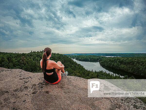 Woman sitting at summit of a hike overlooking lake and trees.