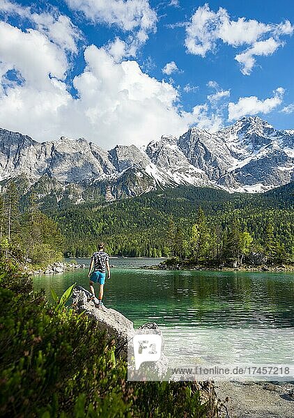 Junger Mann steht auf einem Felsen  Blick in die Ferne  Eibsee und Zugspitze im Frühlingmit Schnee  Wettersteingebirge  bei Grainau  Oberbayern  Bayern  Deutschland  Europa