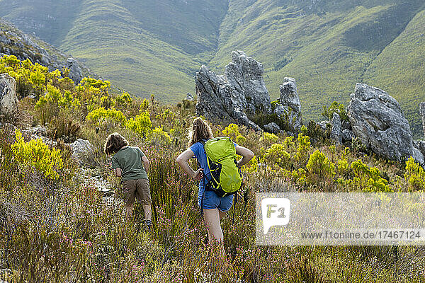 Ein Mädchen und ein Junge wandern auf einem Pfad durch Vegetation und Felsen im Fynbos