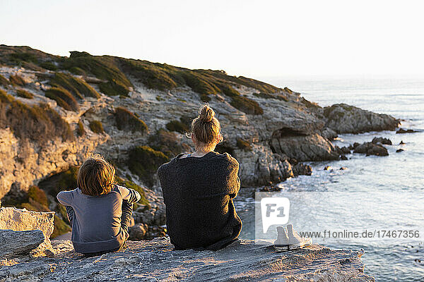 Teenage girl and young boy sitting on rocks looking over the sea at sunset