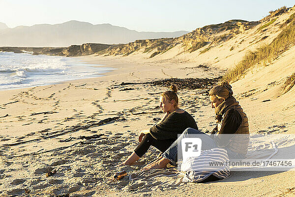 Zwei Personen  eine Mutter und eine Tochter  sitzen auf dem Sand und schauen auf das Meer hinaus