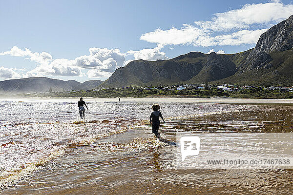 Teenager-Mädchen und ein Junge laufen durch flaches Wasser an einem breiten Sandstrand