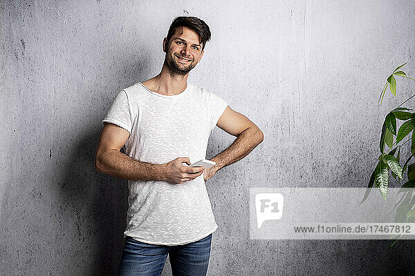Lächelnder Mann mit der Hand auf der Hüfte  der sein Smartphone vor einer weißen Wand hält